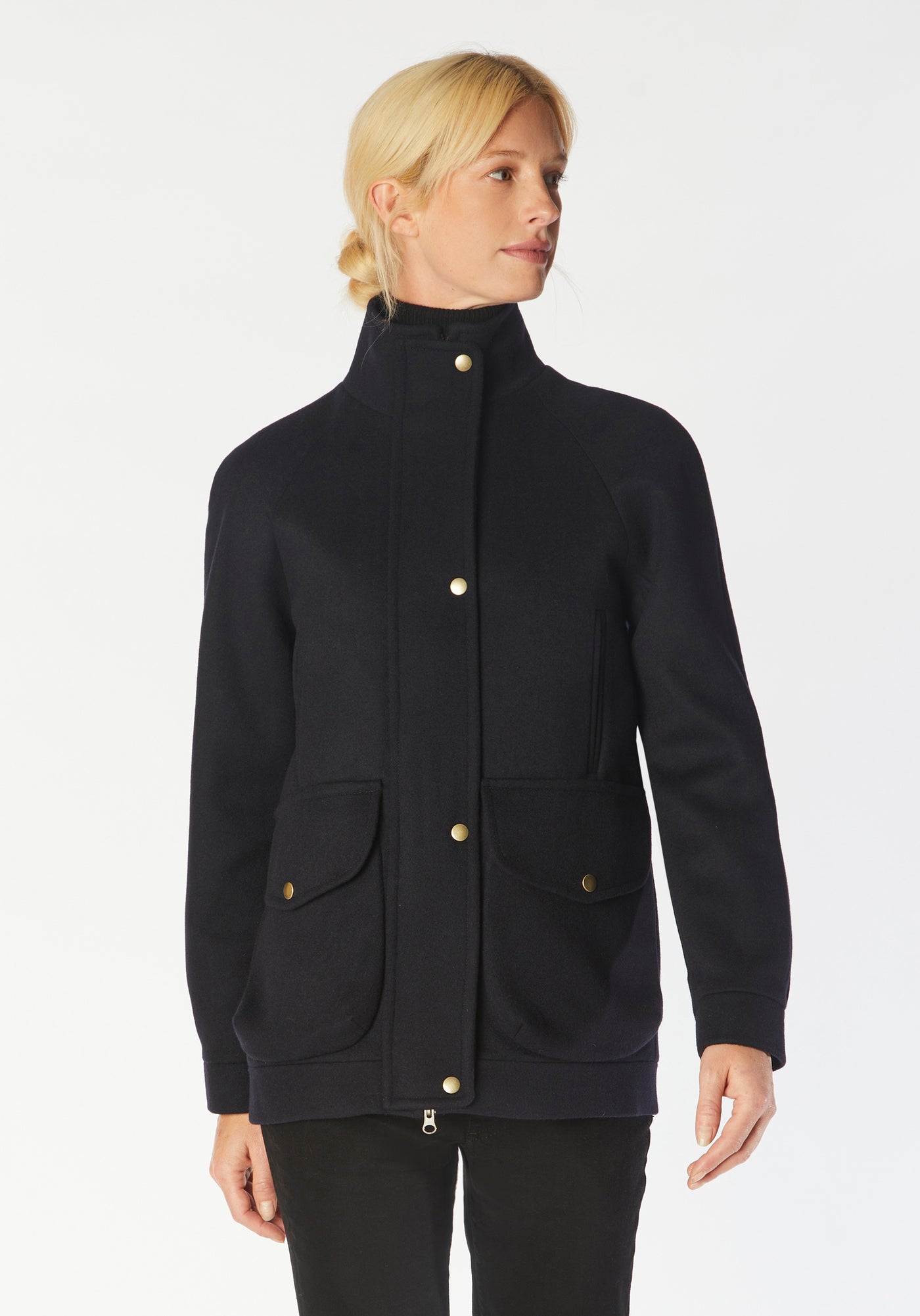A model wears the Andrea jacket in navy merino wool, featuring relaxed raglan sleeves, a high neck, generous pockets, and finished with antique brass hardware.