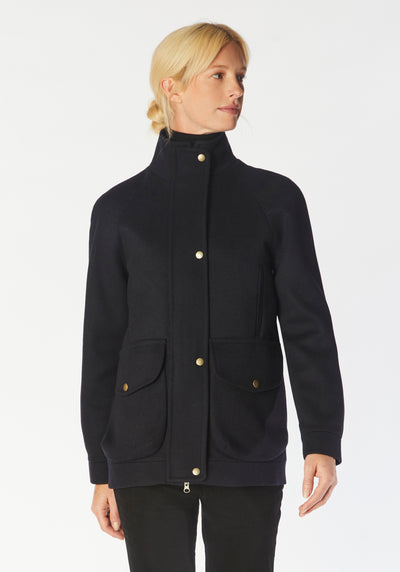 A model wears the Andrea jacket in navy merino wool, featuring relaxed raglan sleeves, a high neck, generous pockets, and finished with antique brass hardware.