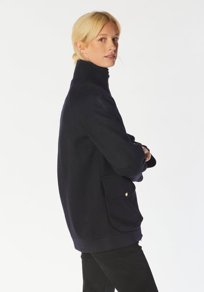 A model wears the Andrea jacket in navy merino wool, featuring relaxed raglan sleeves, a high neck, generous pockets, and finished with antique brass hardware.