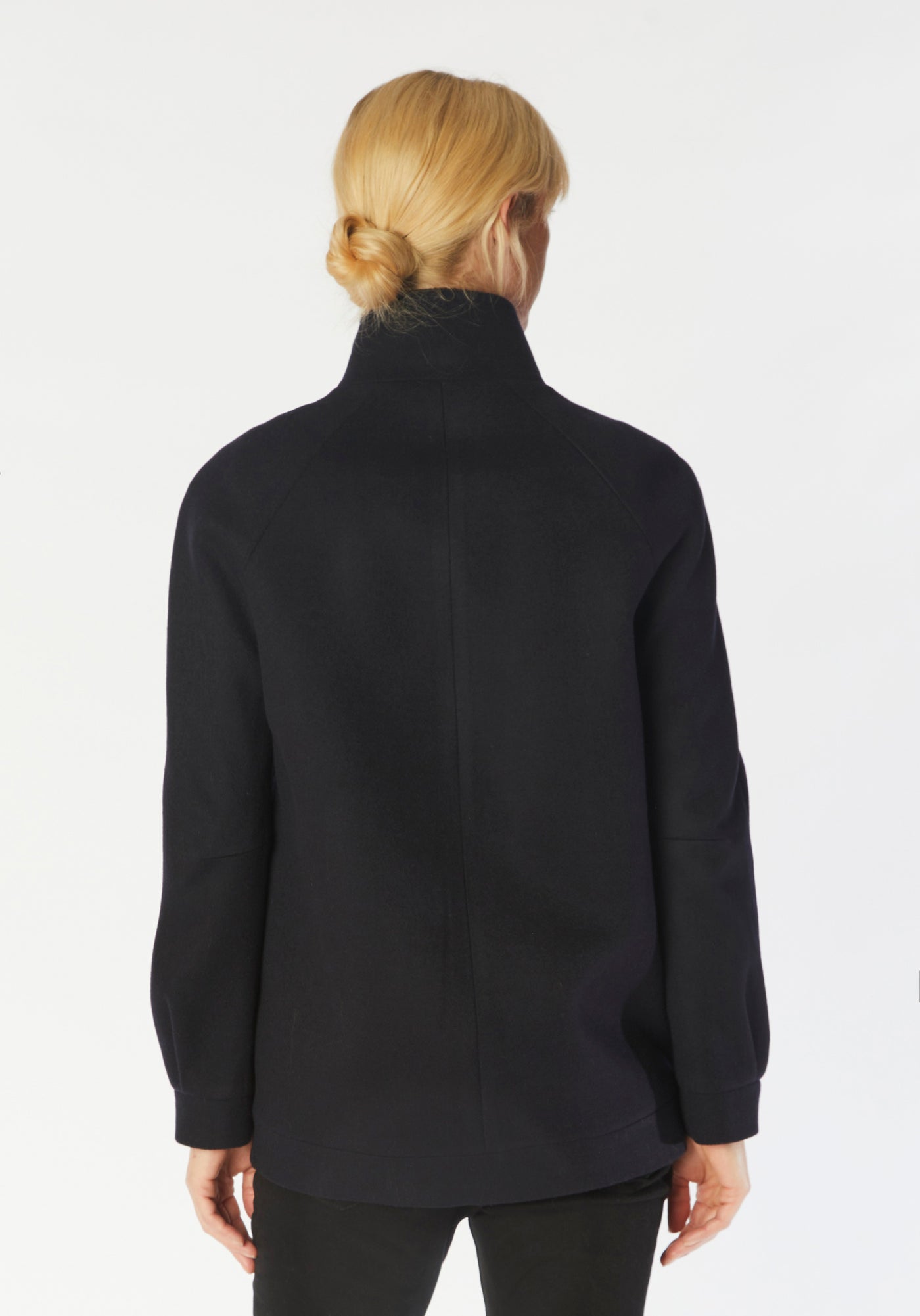 A model wears the Andrea jacket in navy merino wool, featuring relaxed raglan sleeves, a high neck, generous pockets, and finished with antique brass hardware.