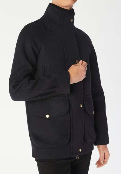 A model wears the Andrea jacket in navy merino wool, featuring relaxed raglan sleeves, a high neck, generous pockets, and finished with antique brass hardware.