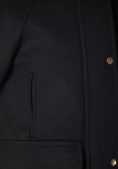 A model wears the Andrea jacket in navy merino wool, featuring relaxed raglan sleeves, a high neck, generous pockets, and finished with antique brass hardware.