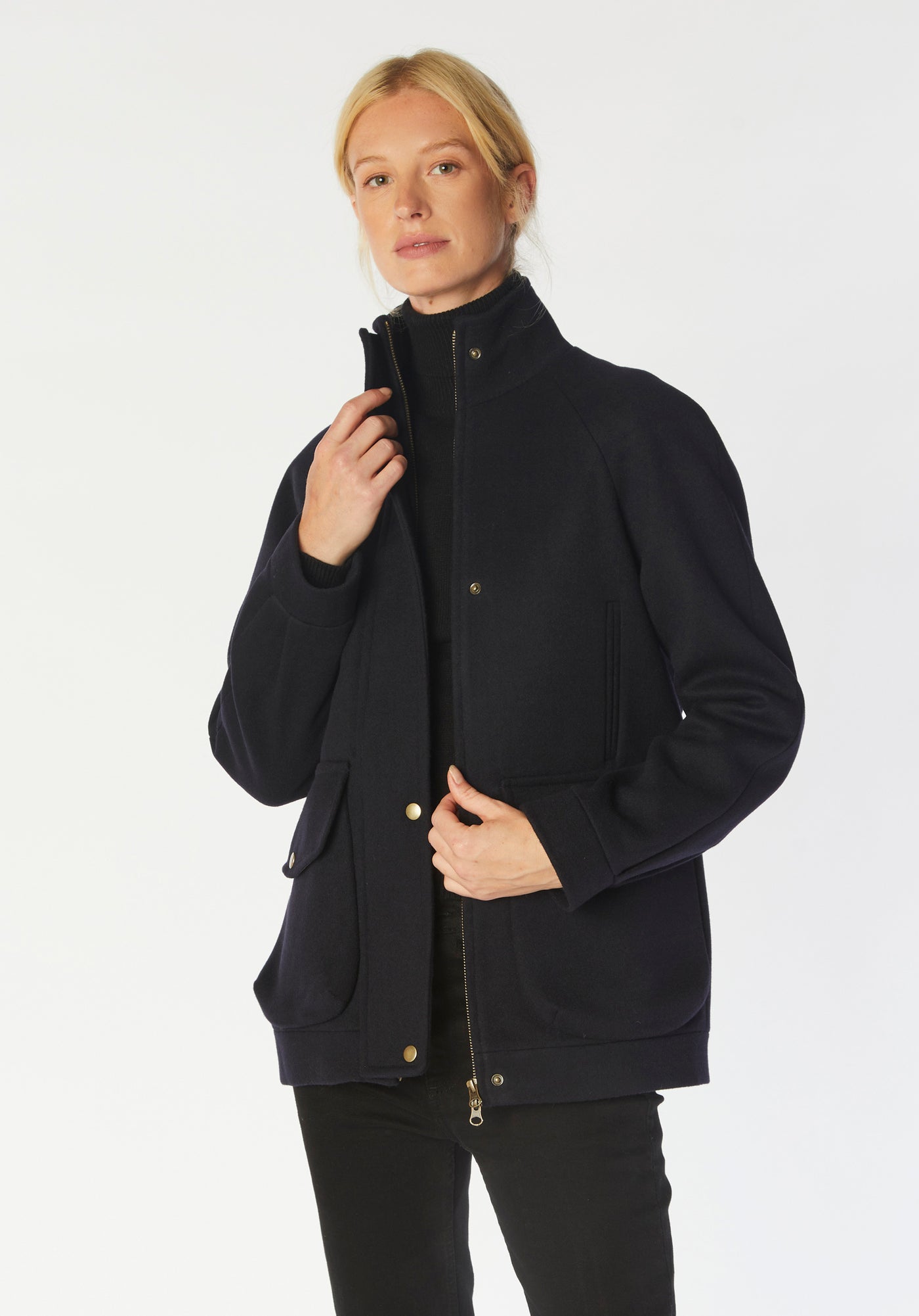 A model wears the Andrea jacket in navy merino wool, featuring relaxed raglan sleeves, a high neck, generous pockets, and finished with antique brass hardware.