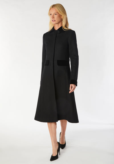 A model wears the Contrast Coat, a long and elegantly tailored black wool cashmere coat with black cotton velvet accents on the collar, cuffs, back belt, and pocket flaps.