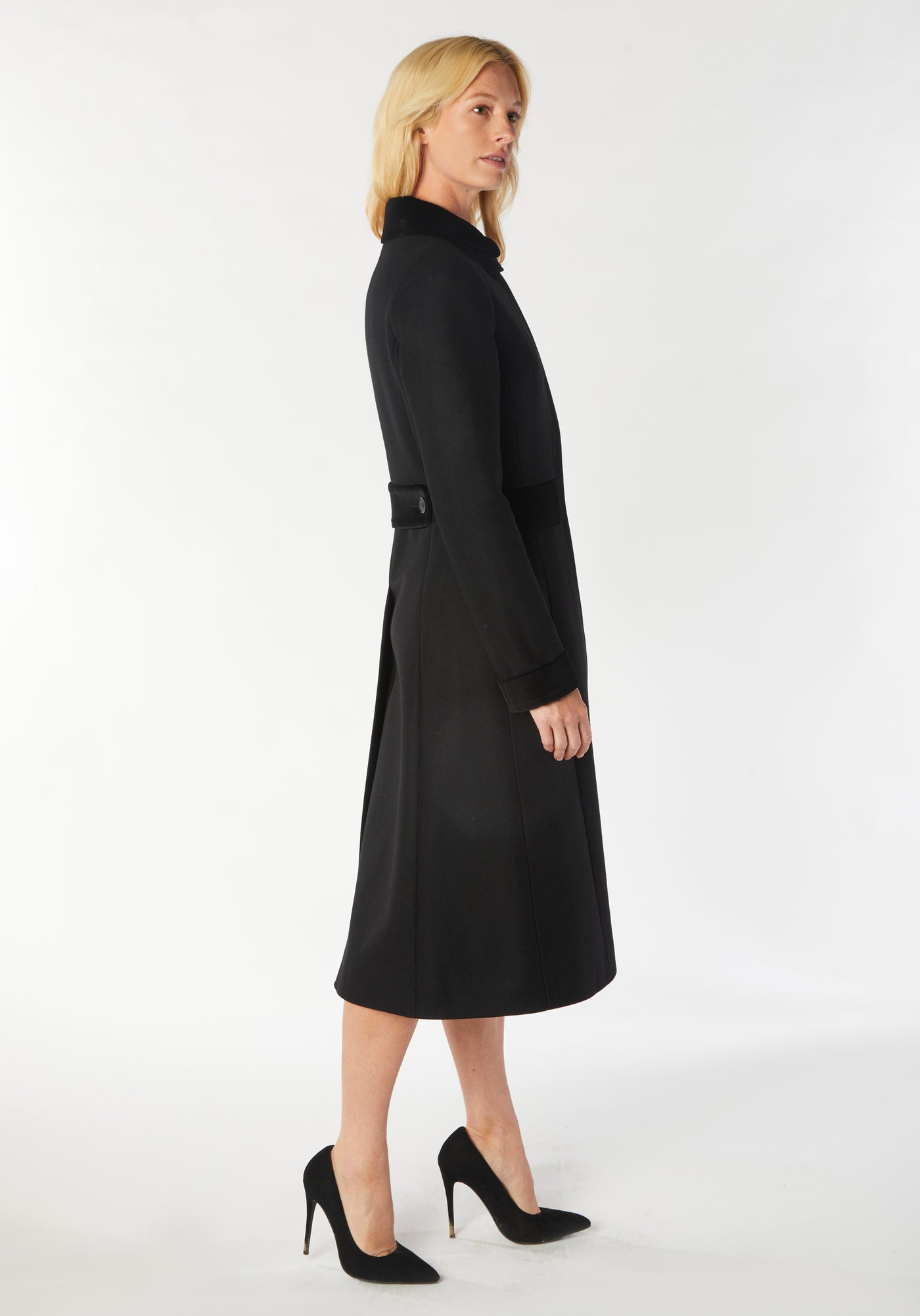 A model wears the Contrast Coat, a long and elegantly tailored black wool cashmere coat with black cotton velvet accents on the collar, cuffs, back belt, and pocket flaps.