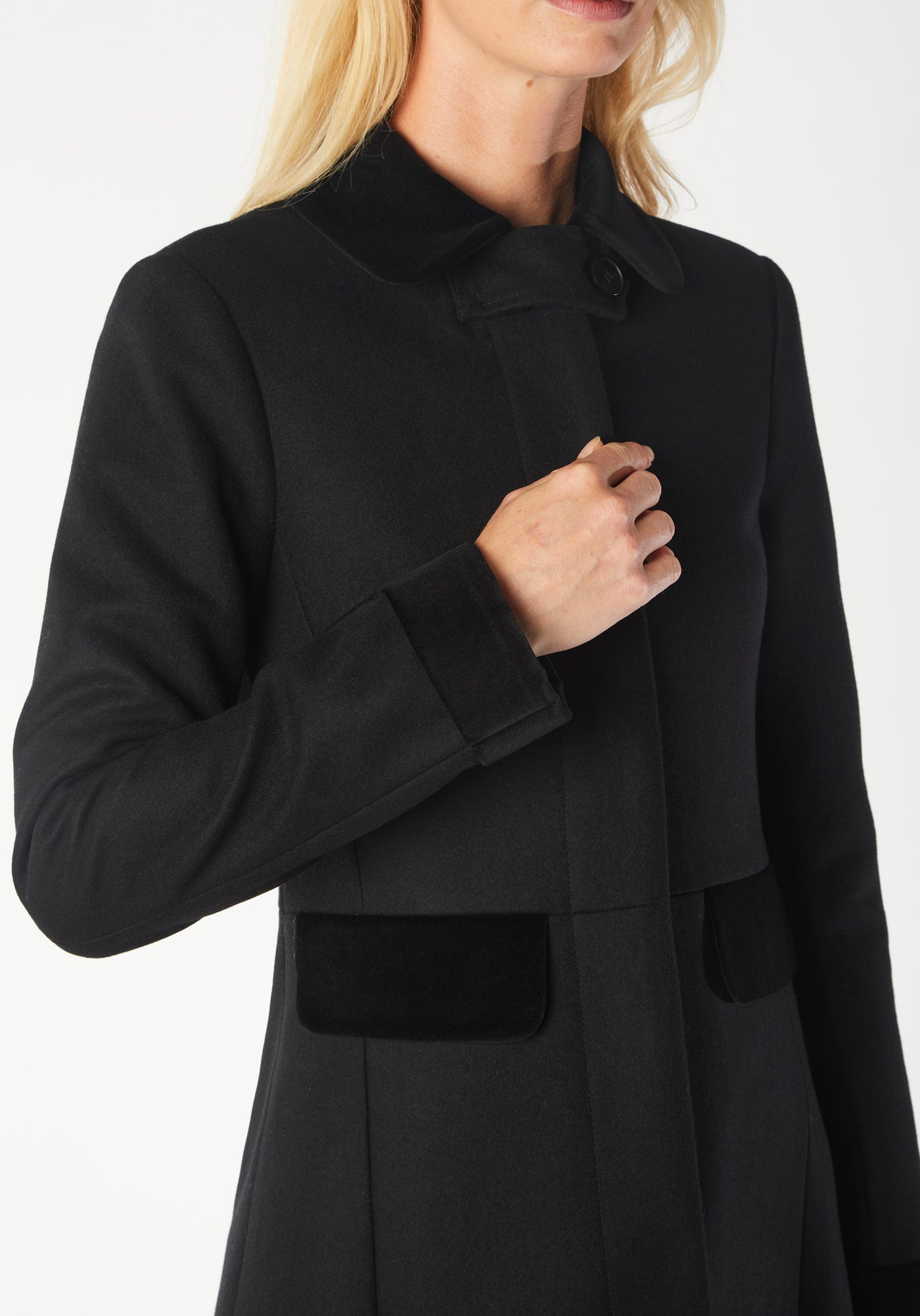 A model wears the Contrast Coat, a long and elegantly tailored black wool cashmere coat with black cotton velvet accents on the collar, cuffs, back belt, and pocket flaps.