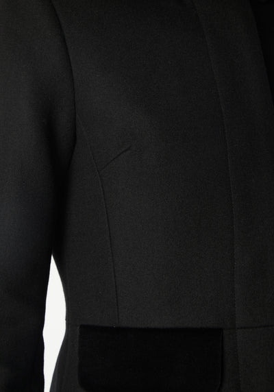 A model wears the Contrast Coat, a long and elegantly tailored black wool cashmere coat with black cotton velvet accents on the collar, cuffs, back belt, and pocket flaps.