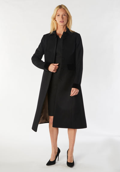 A model wears the Contrast Coat, a long and elegantly tailored black wool cashmere coat with black cotton velvet accents on the collar, cuffs, back belt, and pocket flaps.