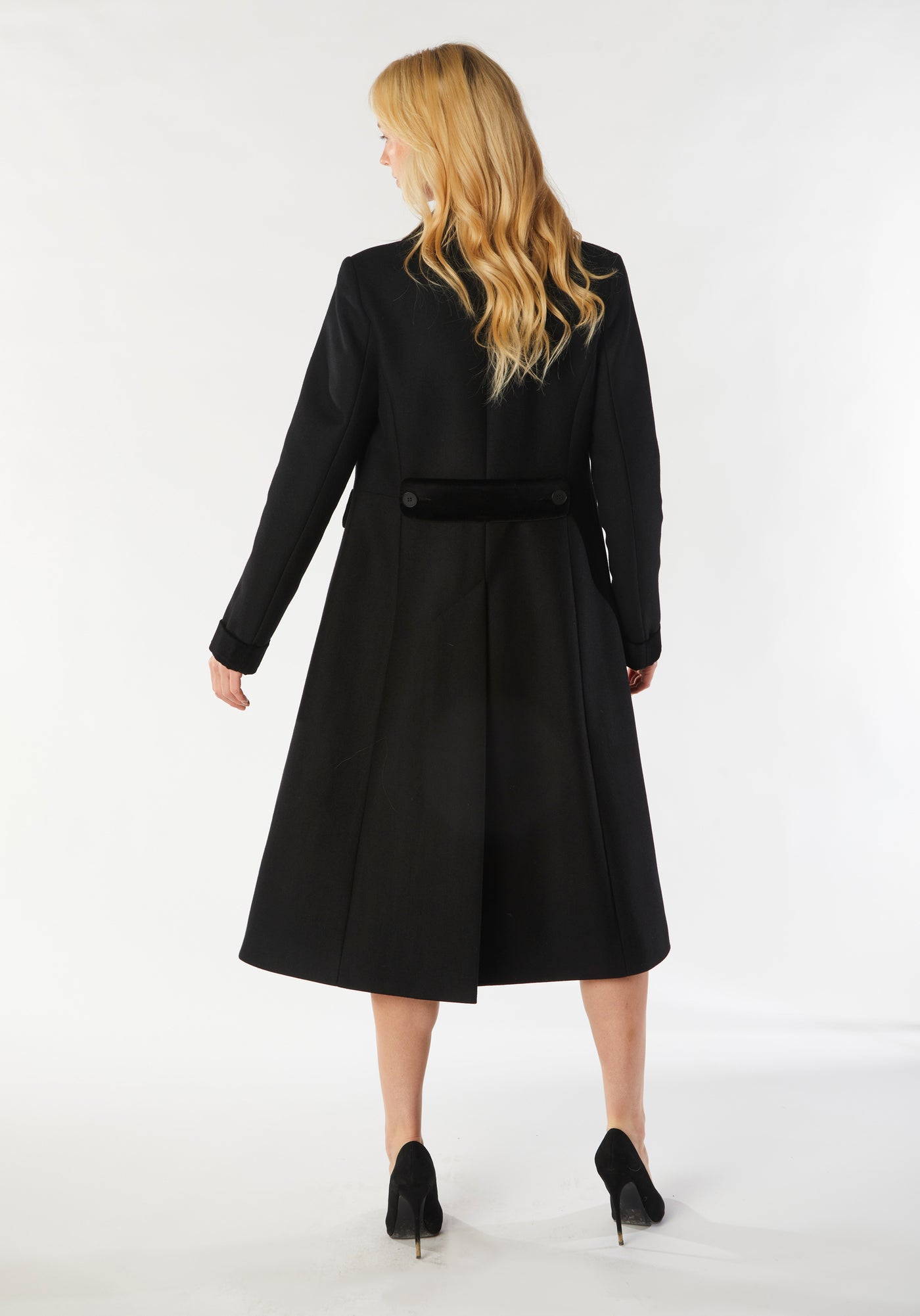 A model wears the Contrast Coat, a long and elegantly tailored black wool cashmere coat with black cotton velvet accents on the collar, cuffs, back belt, and pocket flaps.