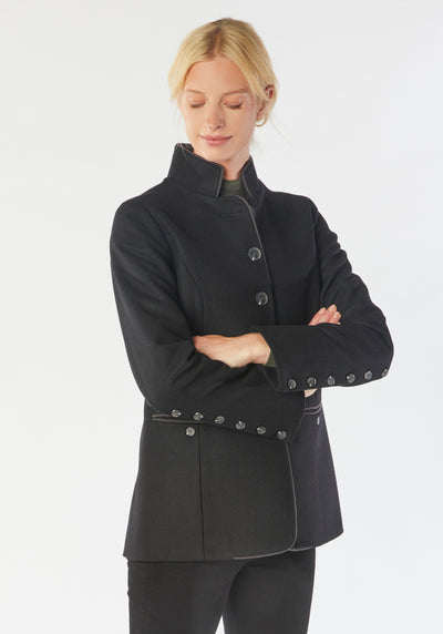 A model wears the Patmos Jacket, a slim-fit design in a black cashmere blend with contrasting grey trim. It features a mandarin collar, front pockets, and deep cuffs with a row of charcoal buttons.