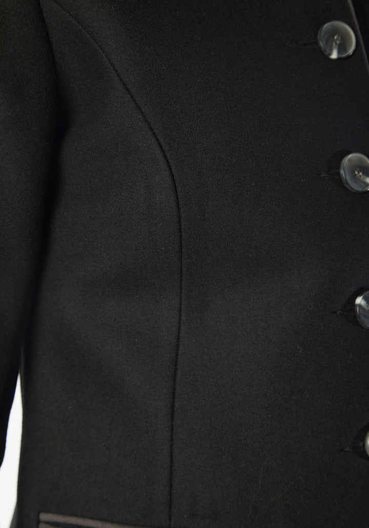 A model wears the Patmos Jacket, a slim-fit design in a black cashmere blend with contrasting grey trim. It features a mandarin collar, front pockets, and deep cuffs with a row of charcoal buttons.