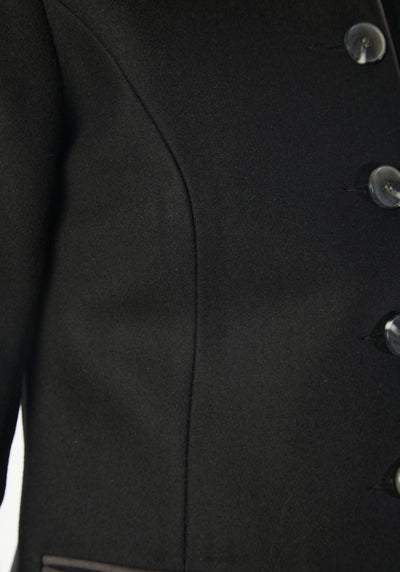 A model wears the Patmos Jacket, a slim-fit design in a black cashmere blend with contrasting grey trim. It features a mandarin collar, front pockets, and deep cuffs with a row of charcoal buttons.