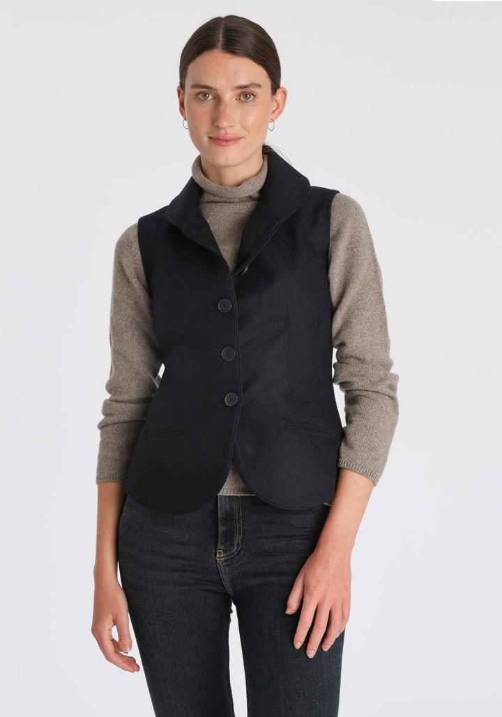 Model wearing the Alexander waistcoat in a navy herringbone lambswool, a short, fitted style with a shawl collar, suede trim, and curved front hem.