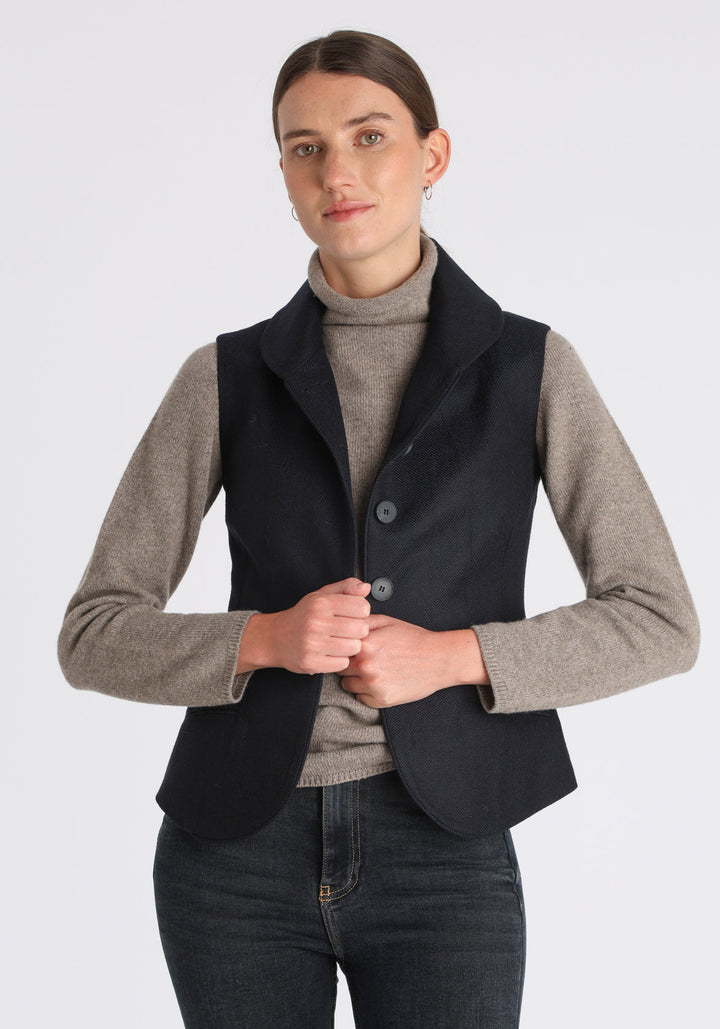Model wearing the Alexander waistcoat in a navy herringbone lambswool, a short, fitted style with a shawl collar, suede trim, and curved front hem.