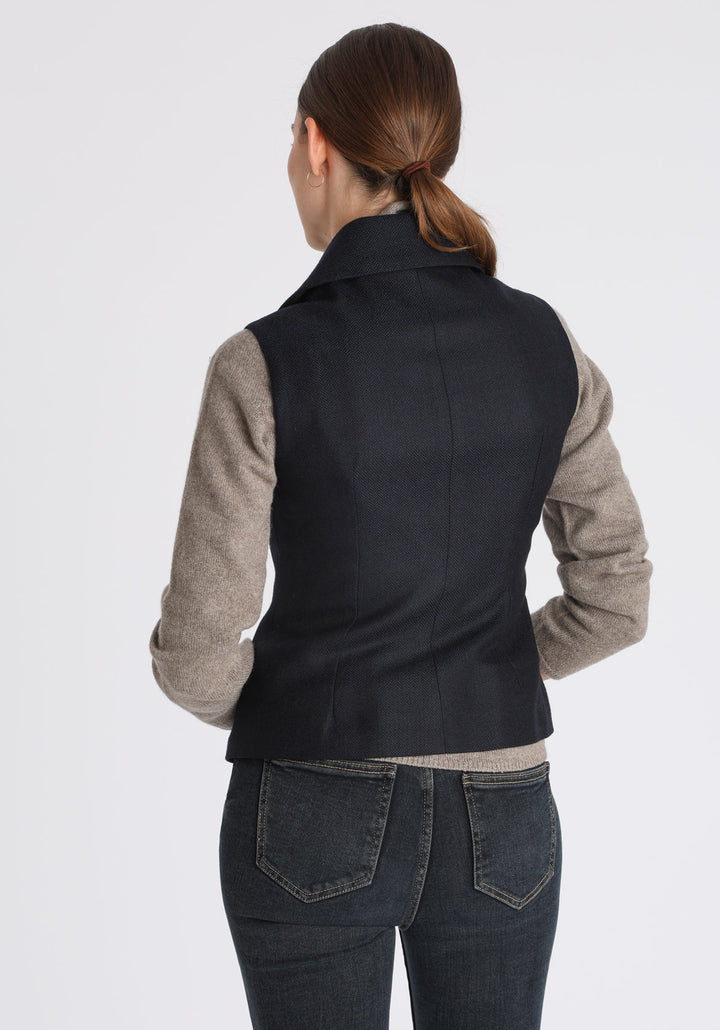 Model wearing the Alexander waistcoat in a navy herringbone lambswool, a short, fitted style with a shawl collar, suede trim, and curved front hem.