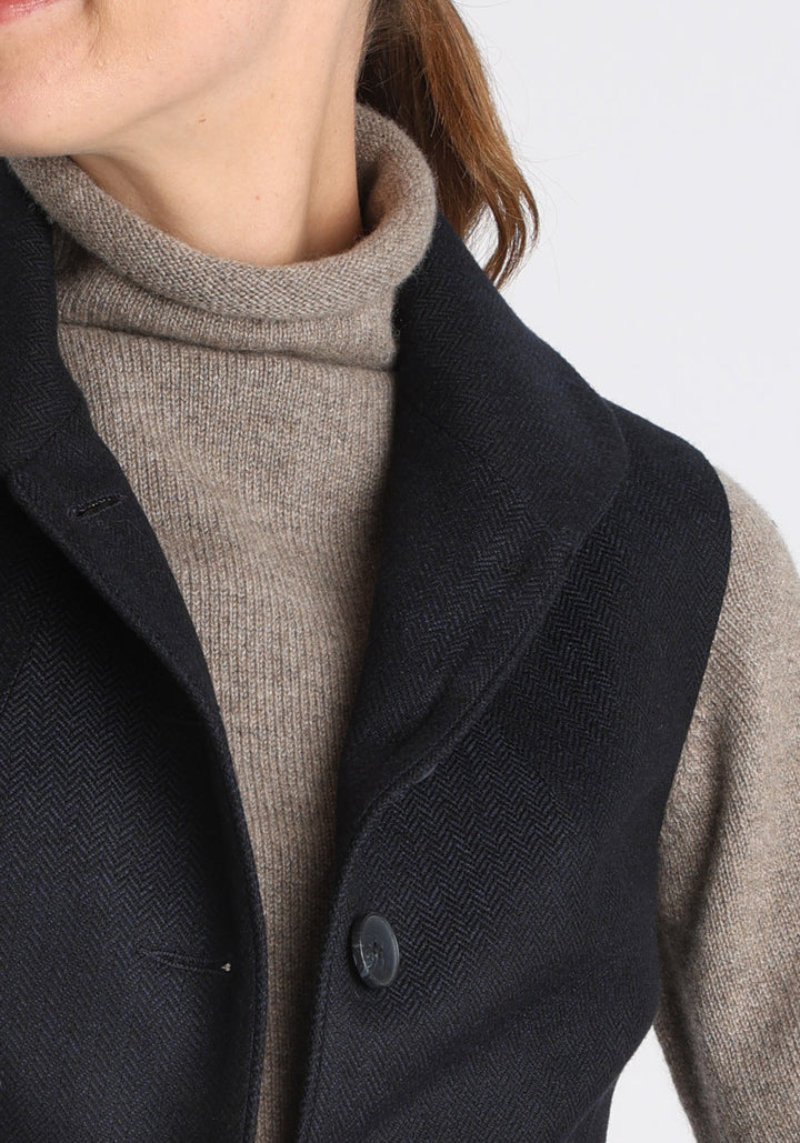 Model wearing the Alexander waistcoat in a navy herringbone lambswool, a short, fitted style with a shawl collar, suede trim, and curved front hem.