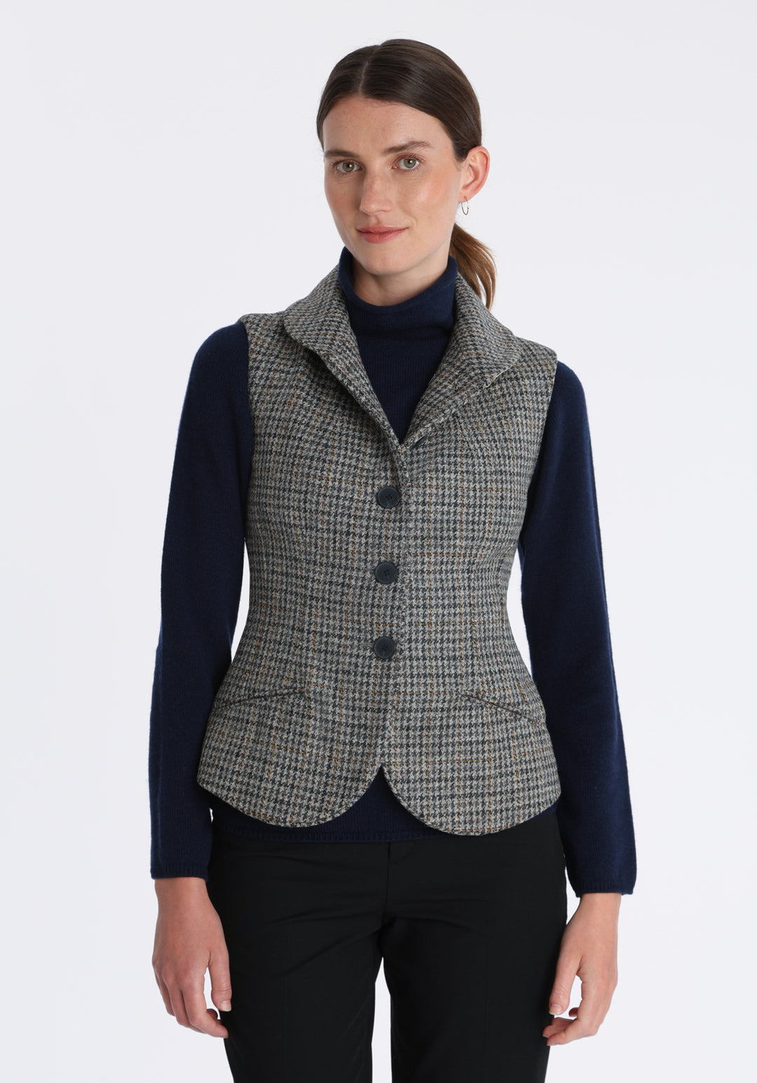 Model wearing the Alexander waistcoat in a stone grey and ochre dogtooth check wool, a short, fitted style with a shawl collar, silk velvet trim, and curved front hem.