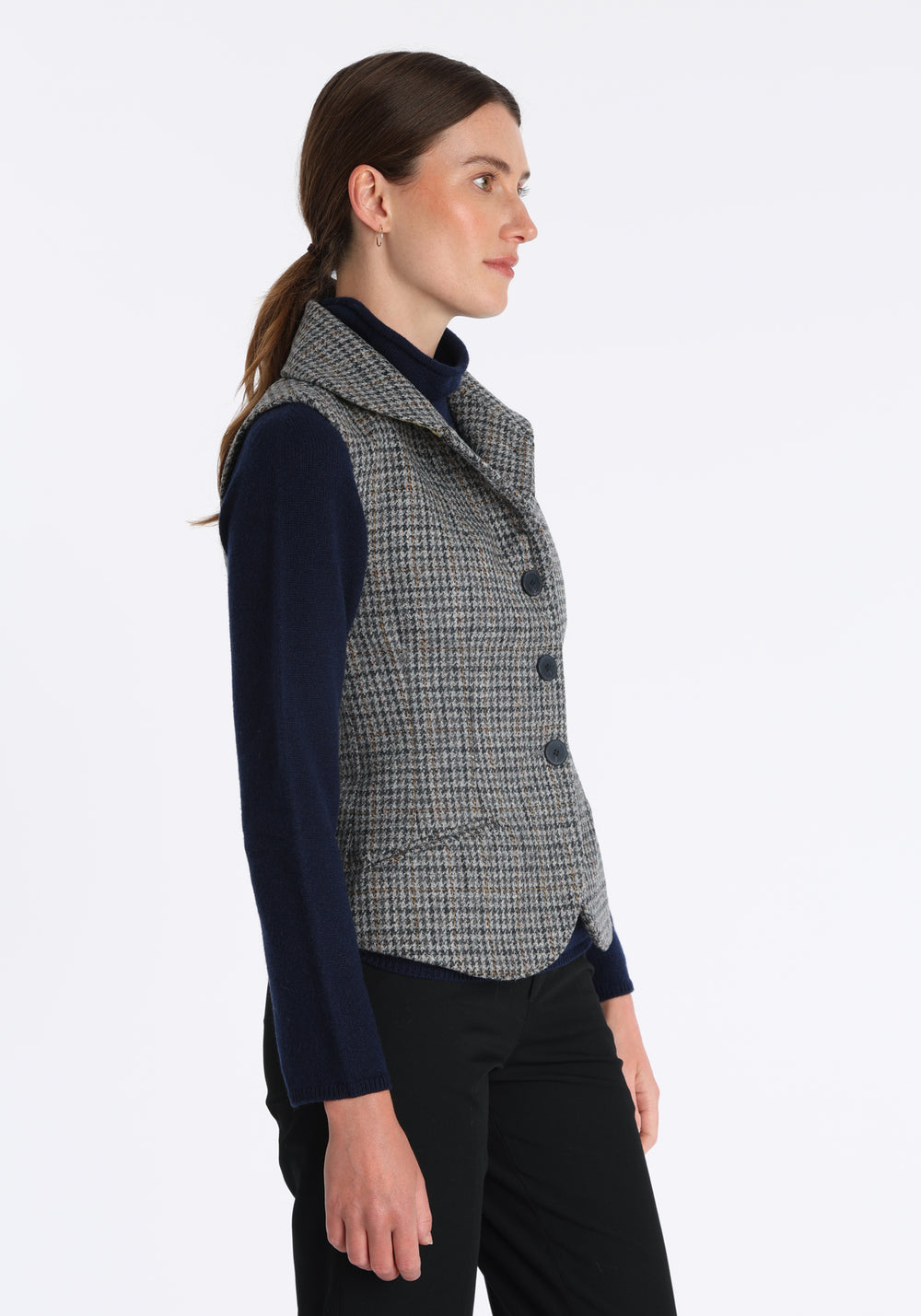 Model wearing the Alexander waistcoat in a stone grey and ochre dogtooth check wool, a short, fitted style with a shawl collar, silk velvet trim, and curved front hem.