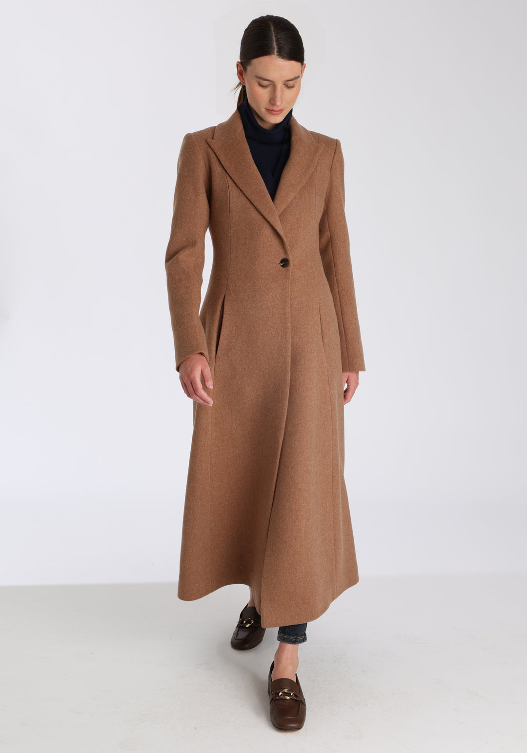 Model wearing the Aris coat in thick camel merino wool, a full-length, double-breasted style with peaked lapels, in-seam pockets, and a cinched waist, designed with a deep back vent for fluid movement.