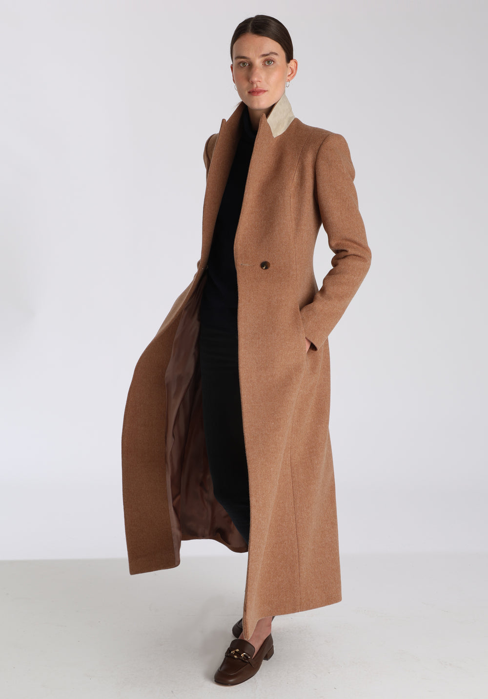 Model wearing the Aris coat in thick camel merino wool, a full-length, double-breasted style with peaked lapels, in-seam pockets, and a cinched waist, designed with a deep back vent for fluid movement.