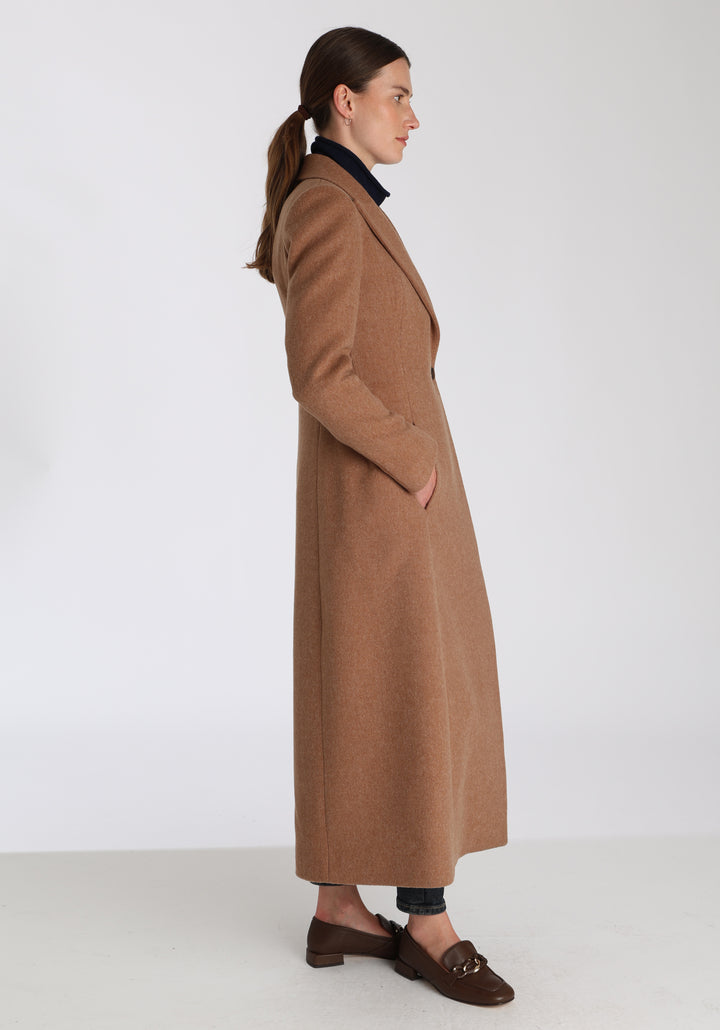 Model wearing the Aris coat in thick camel merino wool, a full-length, double-breasted style with peaked lapels, in-seam pockets, and a cinched waist, designed with a deep back vent for fluid movement.