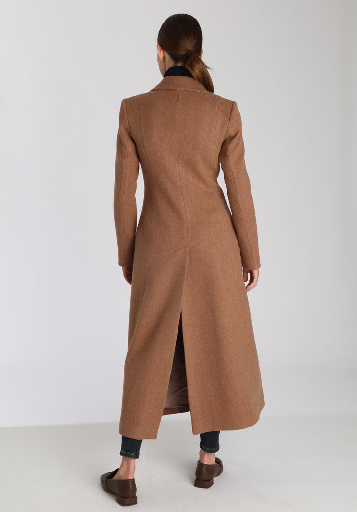 Model wearing the Aris coat in thick camel merino wool, a full-length, double-breasted style with peaked lapels, in-seam pockets, and a cinched waist, designed with a deep back vent for fluid movement.