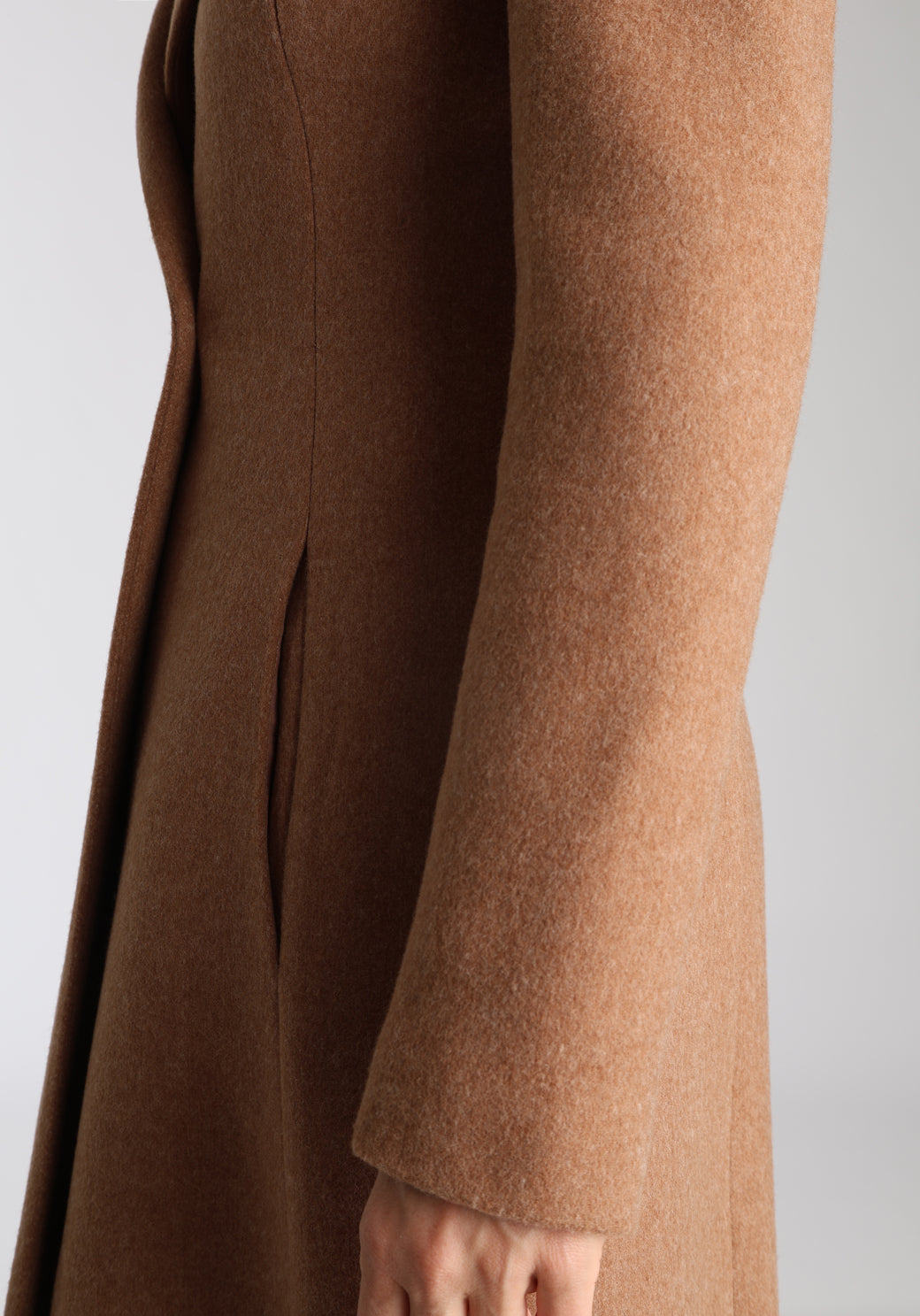 Model wearing the Aris coat in thick camel merino wool, a full-length, double-breasted style with peaked lapels, in-seam pockets, and a cinched waist, designed with a deep back vent for fluid movement.