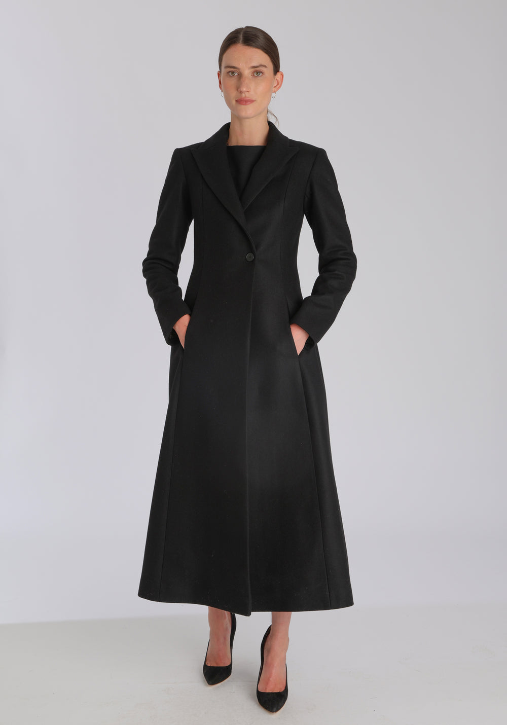 Model wearing the Aris coat in black cashmere blend, a full-length, double-breasted style with peaked lapels, in-seam pockets, and a cinched waist, designed with a deep back vent for fluid movement.