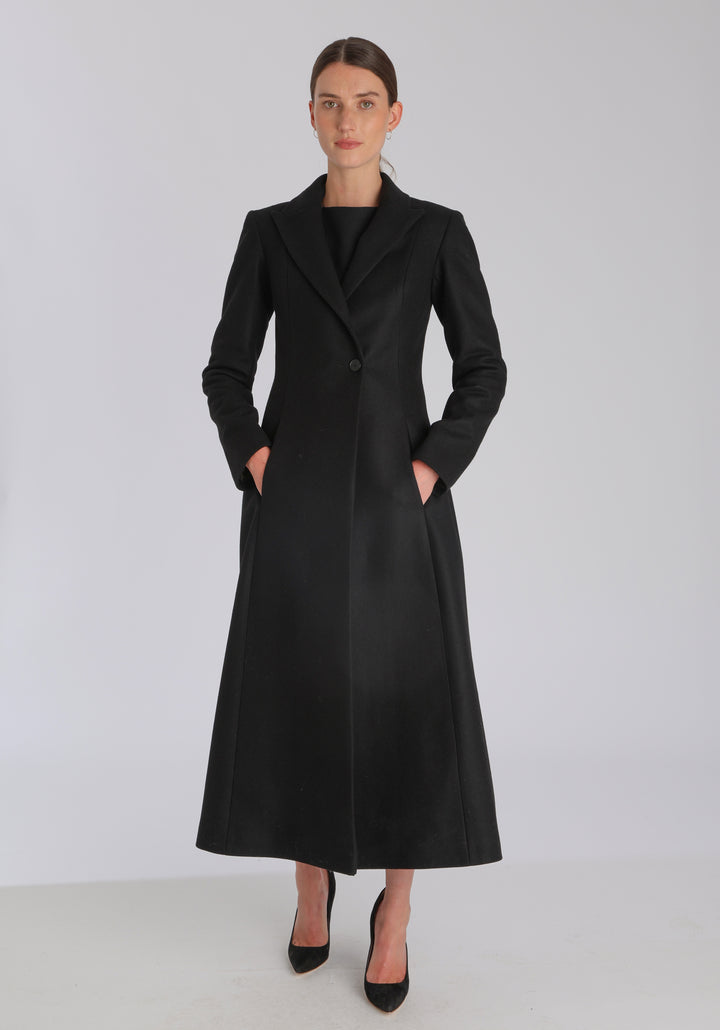 Model wearing the Aris coat in black cashmere blend, a full-length, double-breasted style with peaked lapels, in-seam pockets, and a cinched waist, designed with a deep back vent for fluid movement.