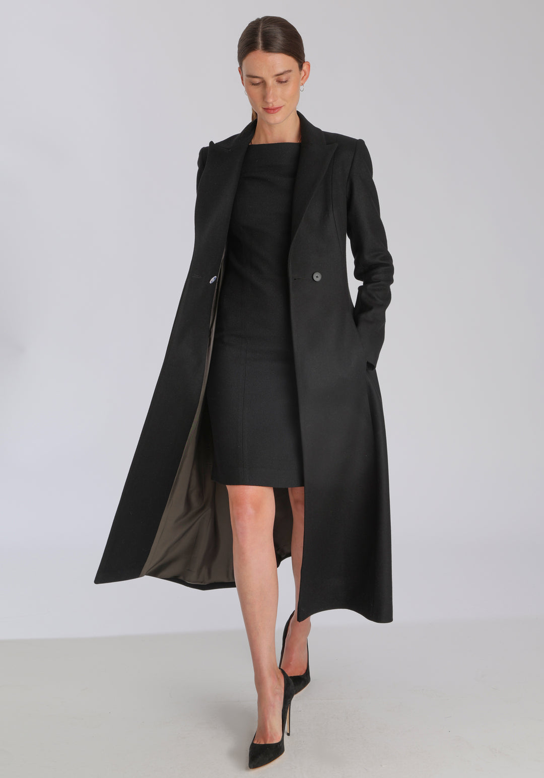 Model wearing the Aris coat in black cashmere blend, a full-length, double-breasted style with peaked lapels, in-seam pockets, and a cinched waist, designed with a deep back vent for fluid movement.