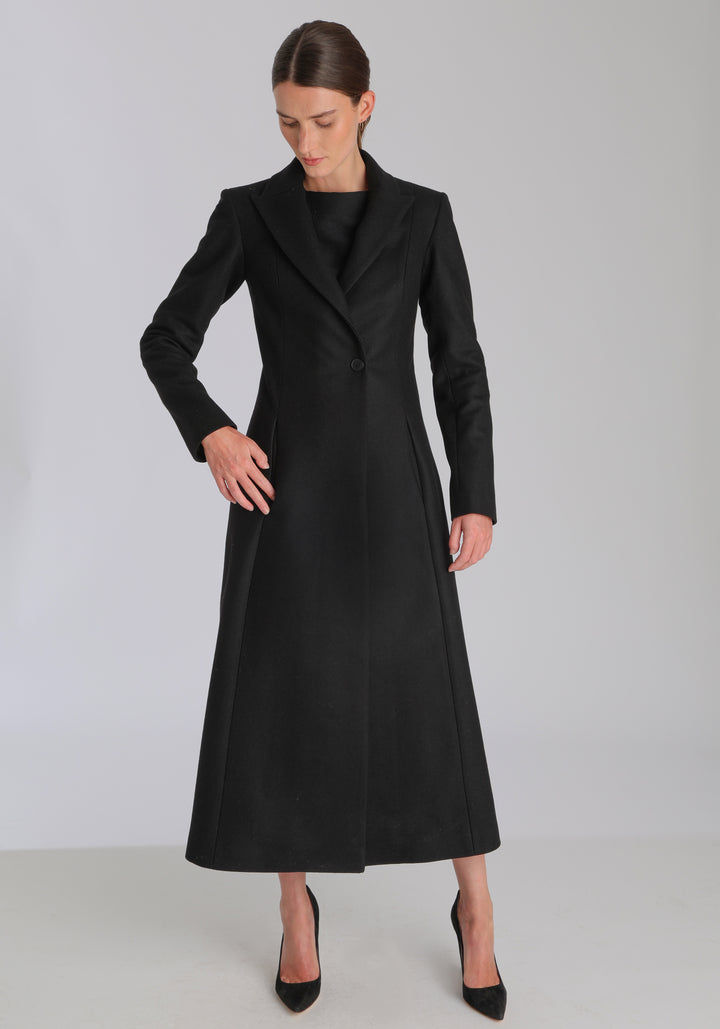 Model wearing the Aris coat in black cashmere blend, a full-length, double-breasted style with peaked lapels, in-seam pockets, and a cinched waist, designed with a deep back vent for fluid movement.