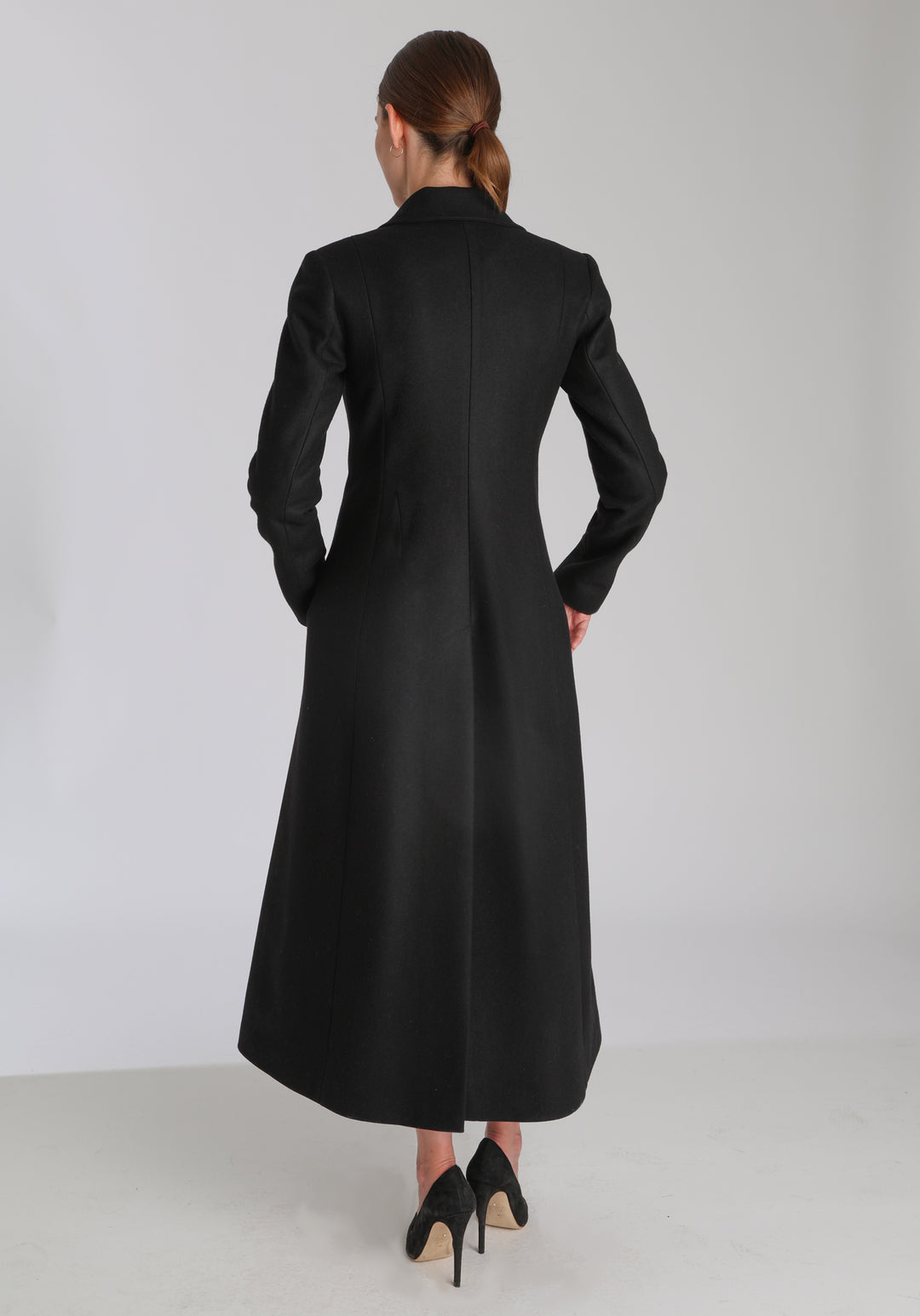 Model wearing the Aris coat in black cashmere blend, a full-length, double-breasted style with peaked lapels, in-seam pockets, and a cinched waist, designed with a deep back vent for fluid movement.
