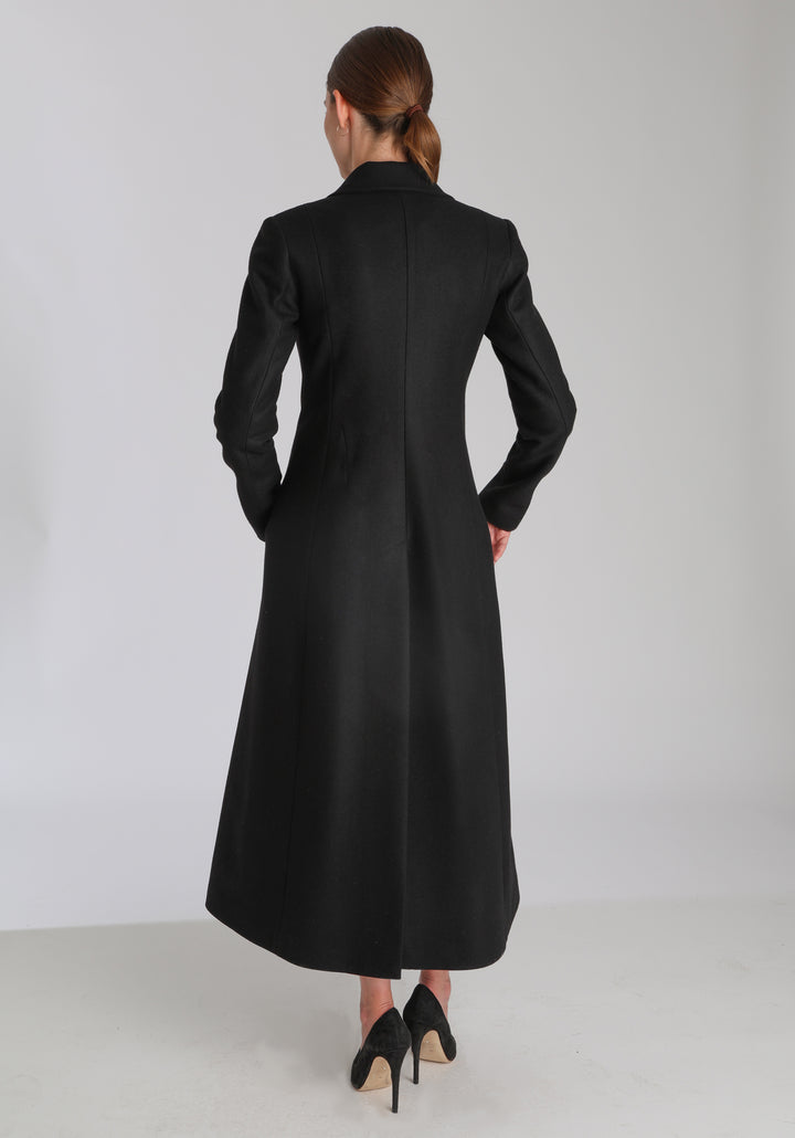 Model wearing the Aris coat in black cashmere blend, a full-length, double-breasted style with peaked lapels, in-seam pockets, and a cinched waist, designed with a deep back vent for fluid movement.
