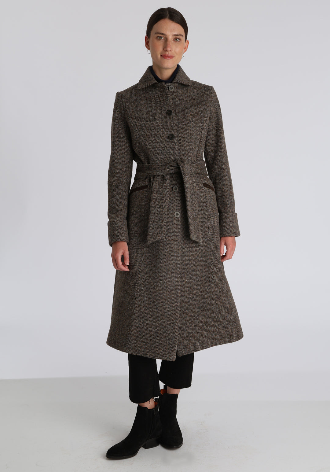 Model wearing the Belted coat in heritage heather herringbone Harris Tweed, a long tailored style with a matching belt, turn-back cuffs, and a deep back vent for ease of movement.