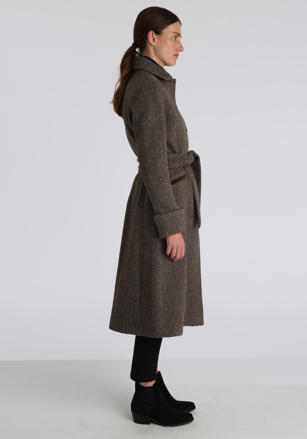 Model wearing the Belted coat in heritage heather herringbone Harris Tweed, a long tailored style with a matching belt, turn-back cuffs, and a deep back vent for ease of movement.