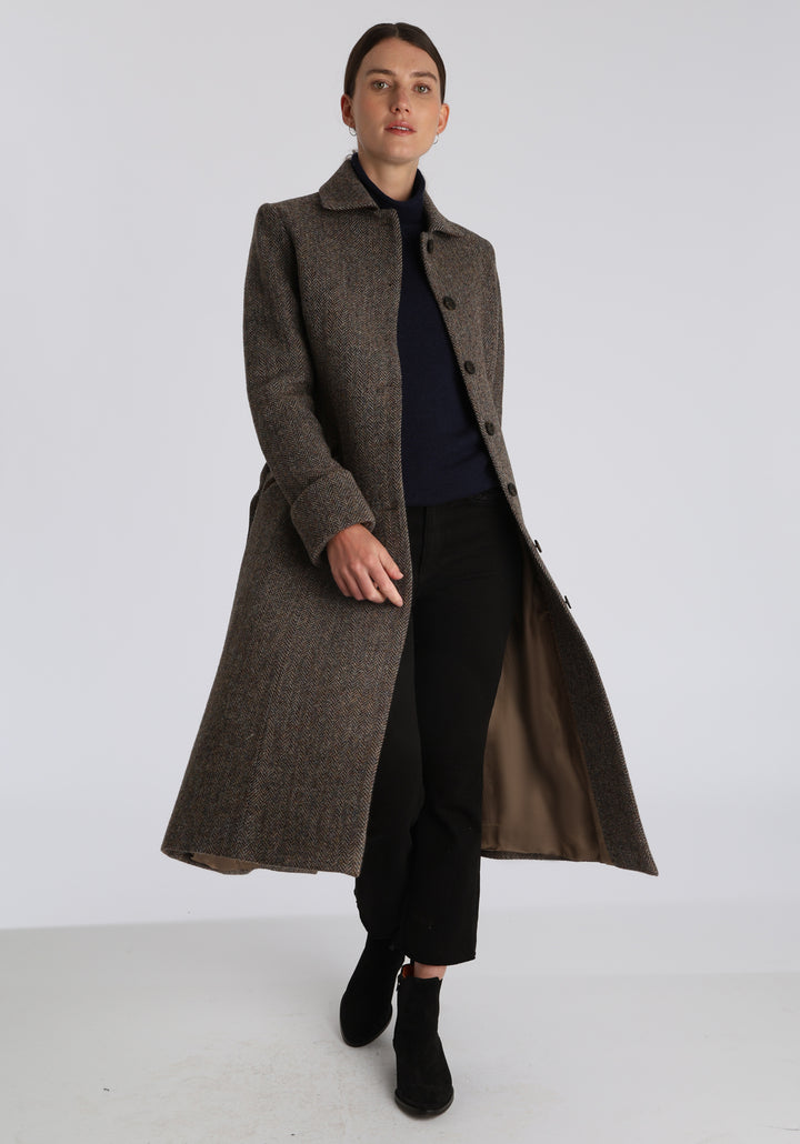 Model wearing the Belted coat in heritage heather herringbone Harris Tweed, a long tailored style with a matching belt, turn-back cuffs, and a deep back vent for ease of movement.