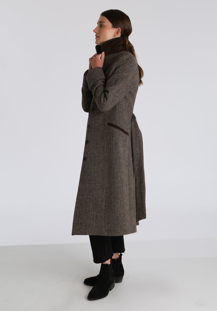 Model wearing the Belted coat in heritage heather herringbone Harris Tweed, a long tailored style with a matching belt, turn-back cuffs, and a deep back vent for ease of movement.