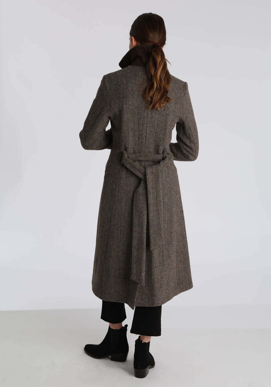 Model wearing the Belted coat in heritage heather herringbone Harris Tweed, a long tailored style with a matching belt, turn-back cuffs, and a deep back vent for ease of movement.