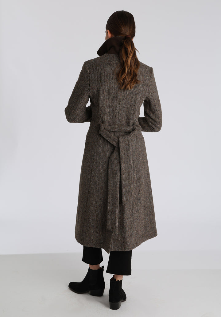 Model wearing the Belted coat in heritage heather herringbone Harris Tweed, a long tailored style with a matching belt, turn-back cuffs, and a deep back vent for ease of movement.
