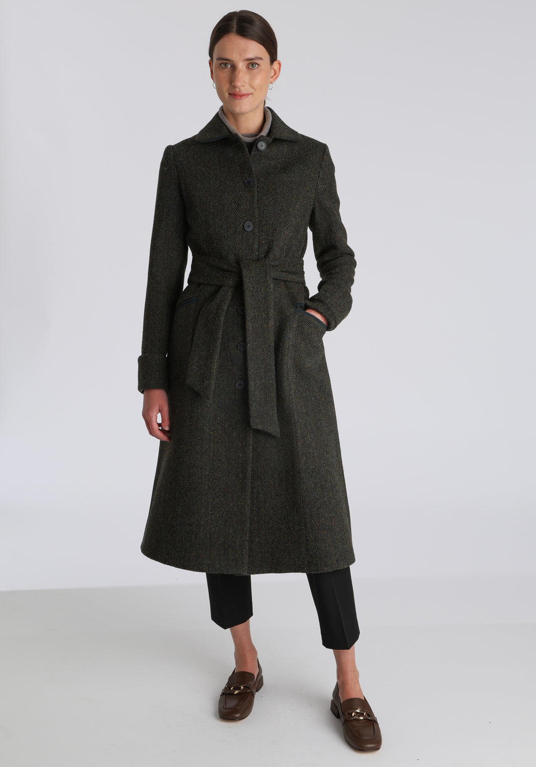 Model wearing the Belted coat in green spruce herringbone Harris Tweed, a long tailored style with a matching belt, turn-back cuffs, and a deep back vent for ease of movement.