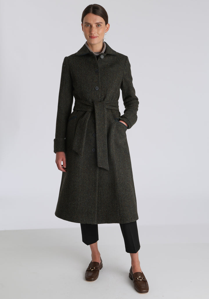 Model wearing the Belted coat in green spruce herringbone Harris Tweed, a long tailored style with a matching belt, turn-back cuffs, and a deep back vent for ease of movement.