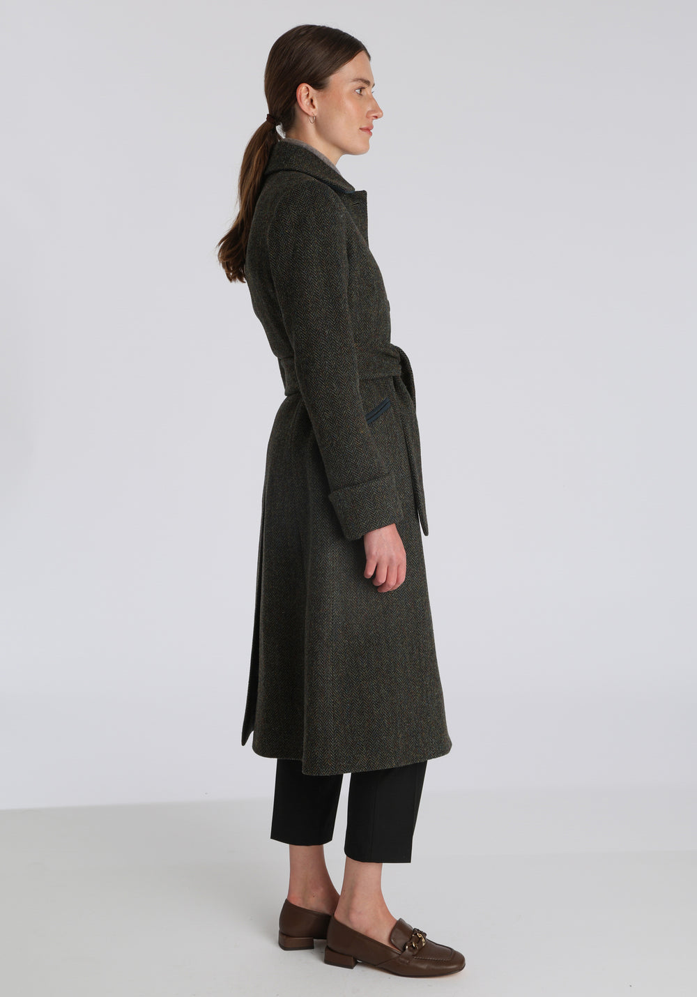 Model wearing the Belted coat in green spruce herringbone Harris Tweed, a long tailored style with a matching belt, turn-back cuffs, and a deep back vent for ease of movement.