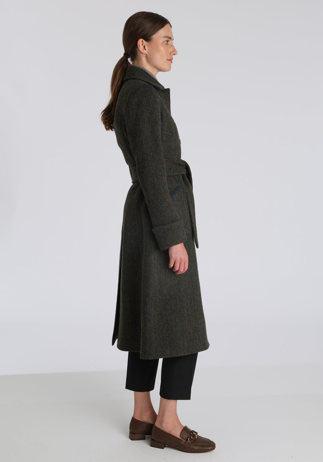 Model wearing the Belted coat in green spruce herringbone Harris Tweed, a long tailored style with a matching belt, turn-back cuffs, and a deep back vent for ease of movement.