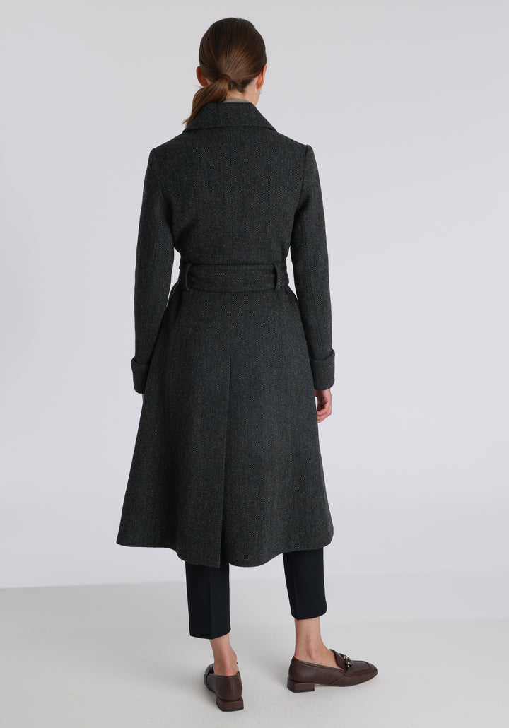 Model wearing the Belted coat in green spruce herringbone Harris Tweed, a long tailored style with a matching belt, turn-back cuffs, and a deep back vent for ease of movement.