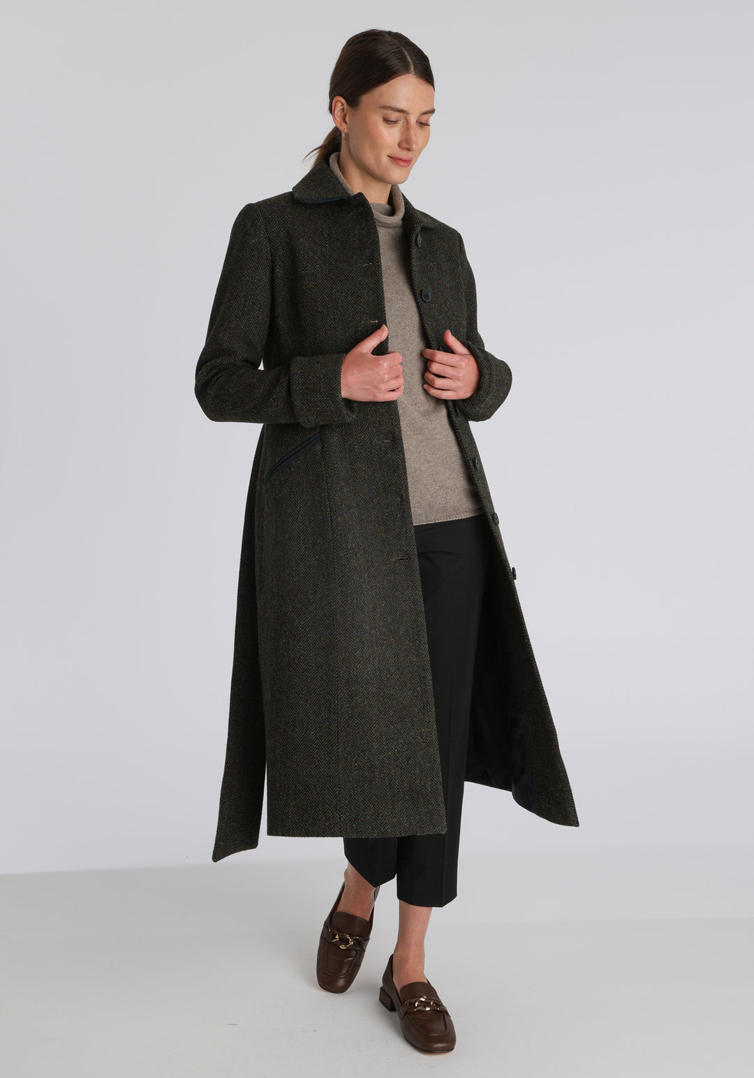 Model wearing the Belted coat in green spruce herringbone Harris Tweed, a long tailored style with a matching belt, turn-back cuffs, and a deep back vent for ease of movement.