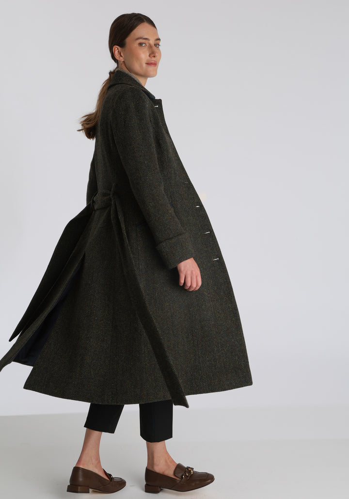 Model wearing the Belted coat in green spruce herringbone Harris Tweed, a long tailored style with a matching belt, turn-back cuffs, and a deep back vent for ease of movement.