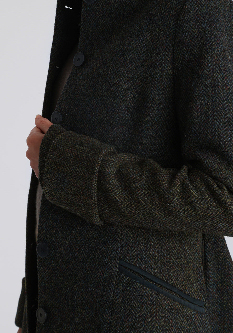 Model wearing the Belted coat in green spruce herringbone Harris Tweed, a long tailored style with a matching belt, turn-back cuffs, and a deep back vent for ease of movement.