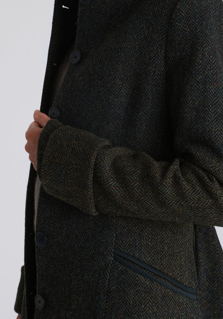 Model wearing the Belted coat in green spruce herringbone Harris Tweed, a long tailored style with a matching belt, turn-back cuffs, and a deep back vent for ease of movement.