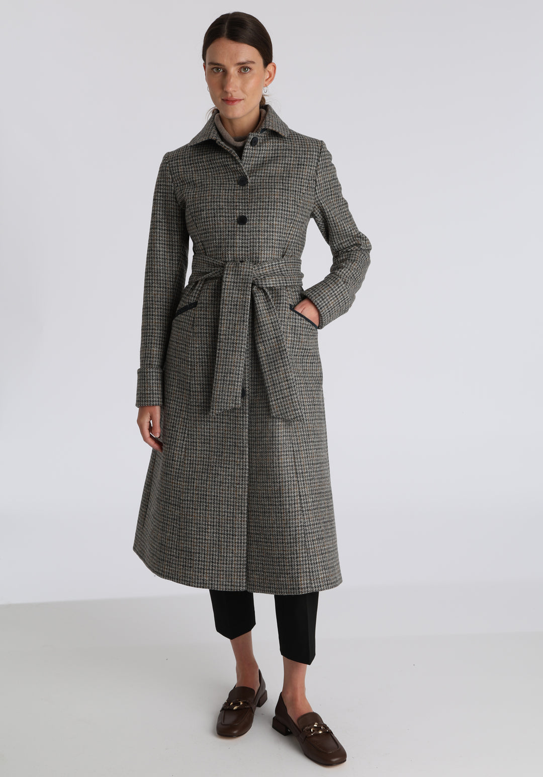 Model wearing the Belted coat in stone grey and ochre dogtooth check wool, a long tailored style with a matching belt, turn-back cuffs, and a deep back vent for ease of movement.