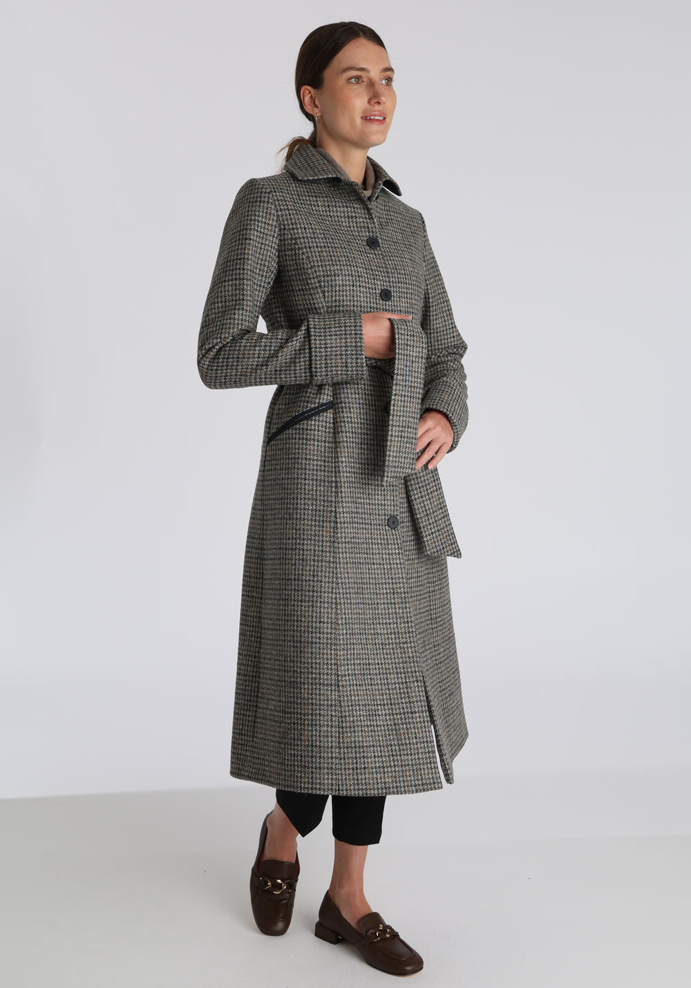 Model wearing the Belted coat in stone grey and ochre dogtooth check wool, a long tailored style with a matching belt, turn-back cuffs, and a deep back vent for ease of movement.